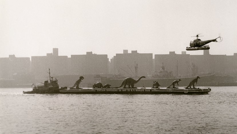 A black and white photograph of a barge carrying dinosaur sculptures on a river, with a helicopter flying above and a city skyline in the hazy background.