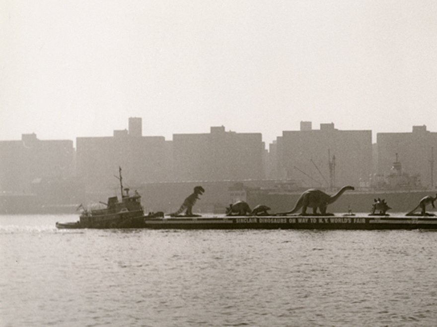 A black and white photograph of a barge carrying dinosaur sculptures on a river, with a helicopter flying above and a city skyline in the hazy background.