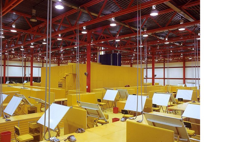 A large, open-plan office space with numerous yellow cubicles and drafting tables under a high ceiling with exposed red beams and bright overhead lighting.