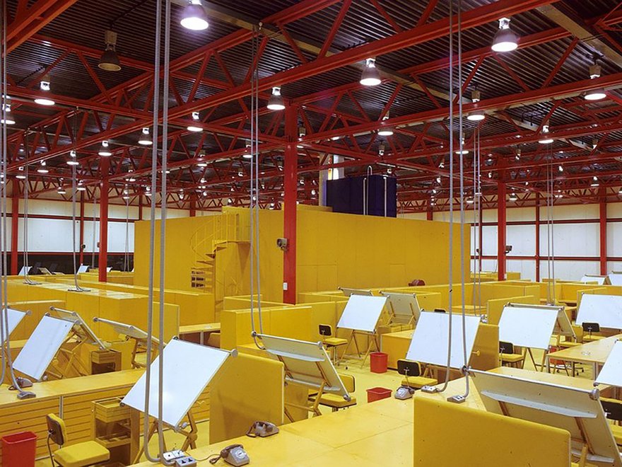 A large, open-plan office space with numerous yellow cubicles and drafting tables under a high ceiling with exposed red beams and bright overhead lighting.