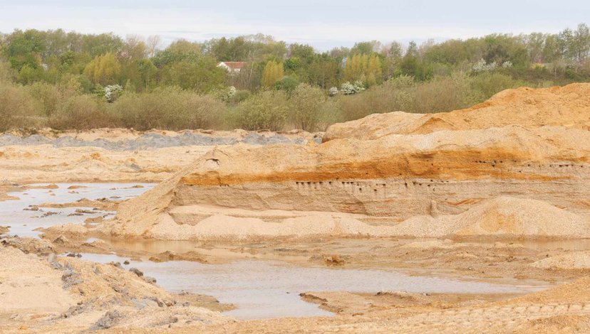 A landscape view of a sand or gravel quarry with large piles of excavated material, some water bodies, and a distant treeline under an overcast sky.