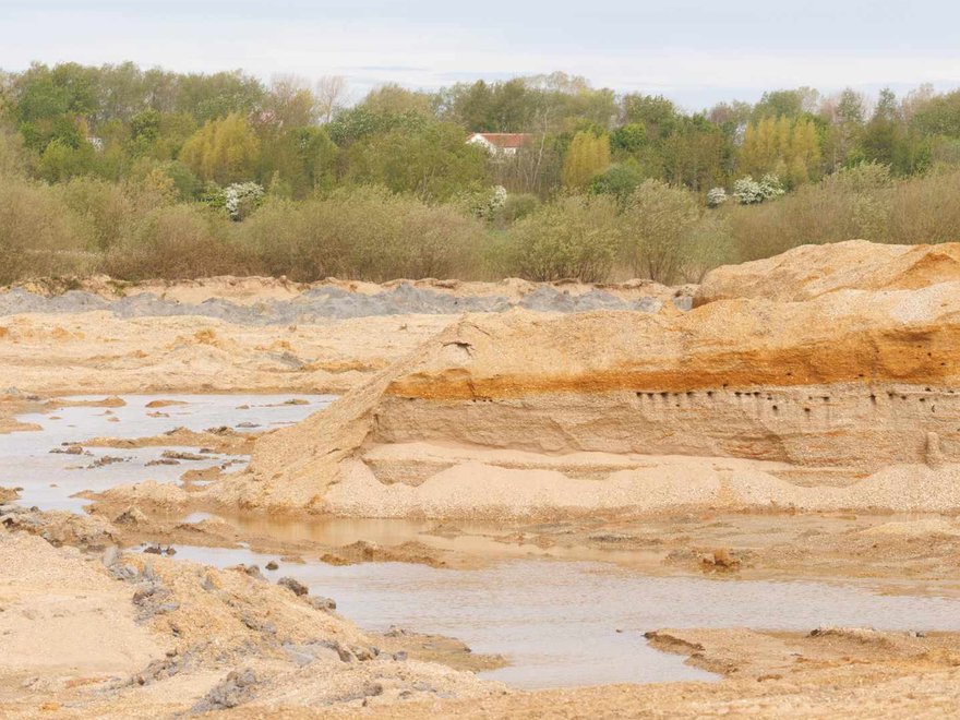 A landscape view of a sand or gravel quarry with large piles of excavated material, some water bodies, and a distant treeline under an overcast sky.