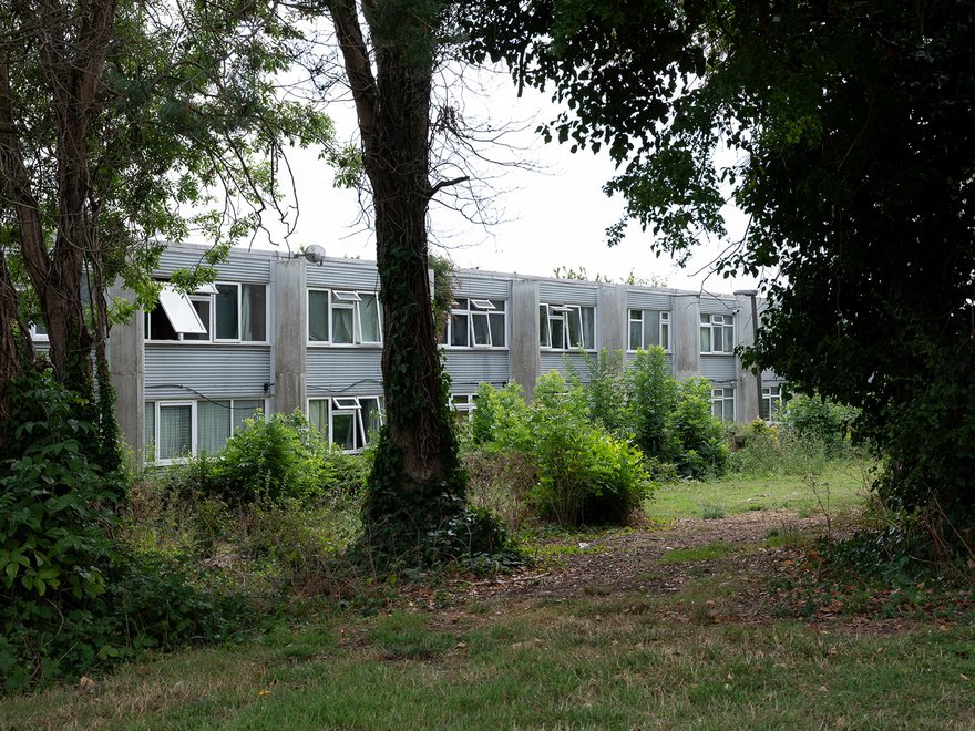 A two-story, light gray building with multiple windows, some open, is partially obscured by trees and overgrown bushes in the foreground and around the structure.
