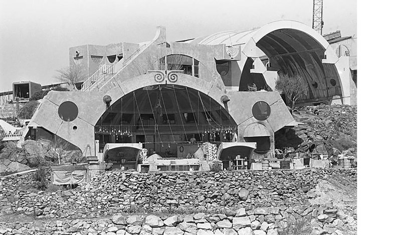 a black and white photograph of Arcosanti, an experimental urban development in the Arizona desert, featuring distinct architectural structures with large arched openings and a surrounding rocky landscape.