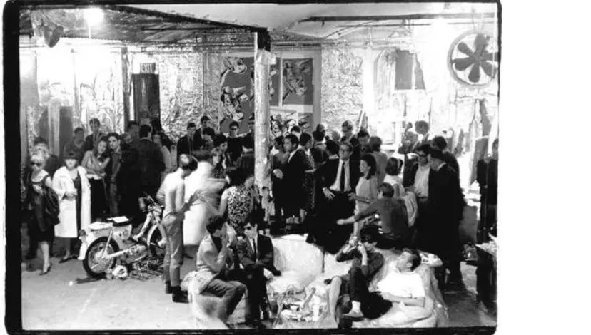 A black and white photograph depicting a crowded gathering of various individuals within what appears to be Andy Warhol's Factory, featuring a diverse group of people socializing, with some seated and others standing.