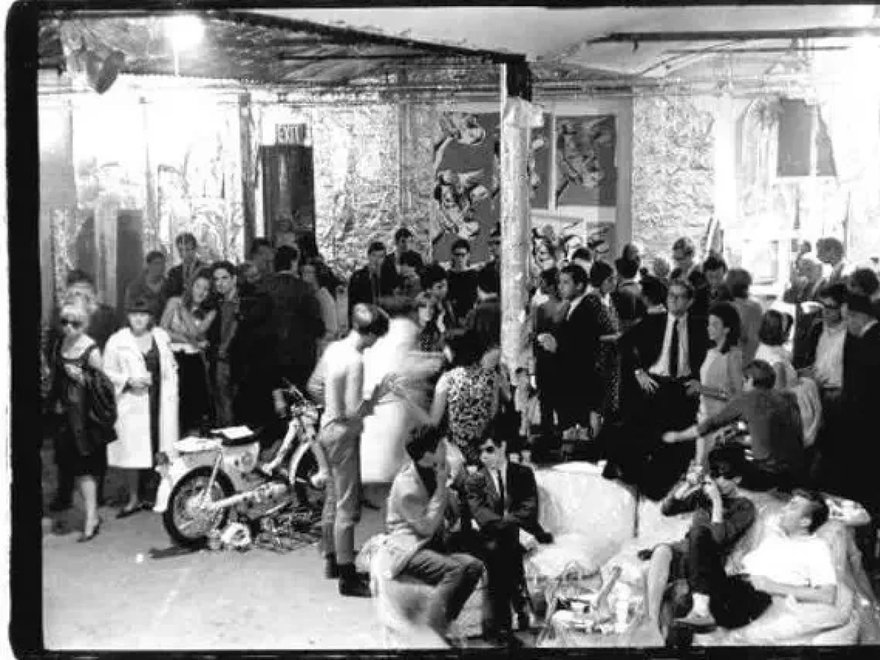 A black and white photograph depicting a crowded gathering of various individuals within what appears to be Andy Warhol's Factory, featuring a diverse group of people socializing, with some seated and others standing.