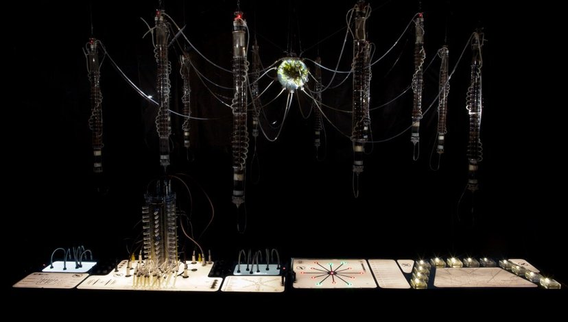 A central glowing, orb-like structure with numerous thin wires extending outwards to several suspended, vertical, cylindrical forms.