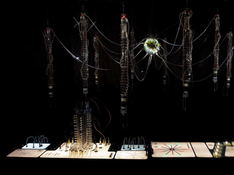 A central glowing, orb-like structure with numerous thin wires extending outwards to several suspended, vertical, cylindrical forms.