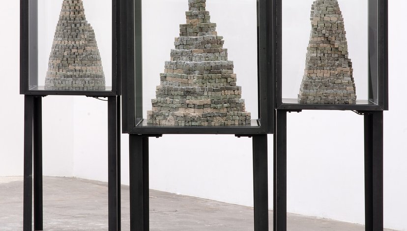 Three tall, narrow display cases, each containing a grey, conical sculpture made of stacked, irregular blocks or bricks.