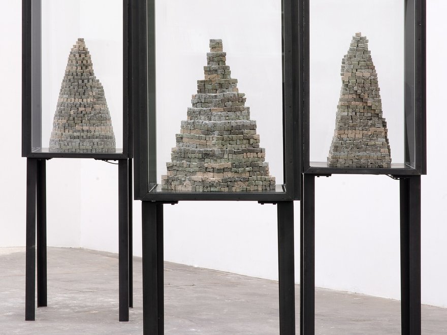 Three tall, narrow display cases, each containing a grey, conical sculpture made of stacked, irregular blocks or bricks.