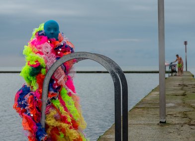 Performer wearing a colourful microbe costume poses in front of the tidal pool in Margate