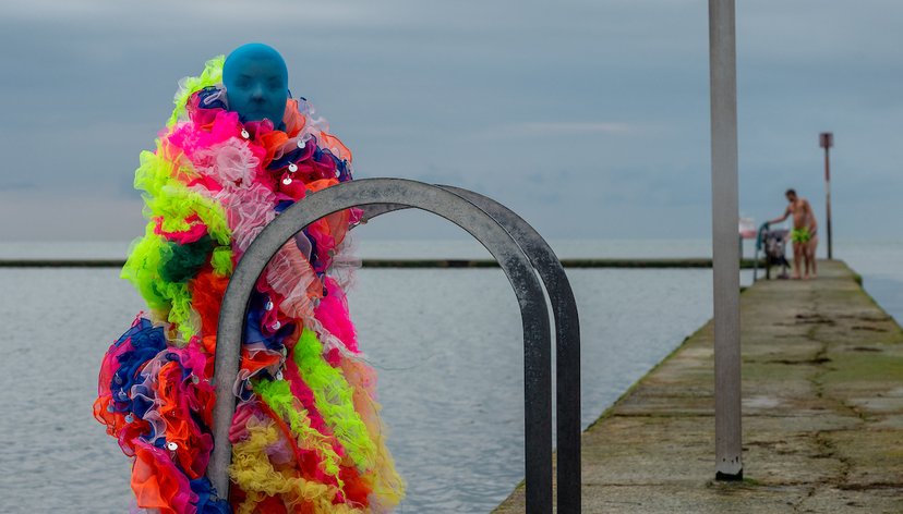 Performer wearing a colourful microbe costume poses in front of the tidal pool in Margate