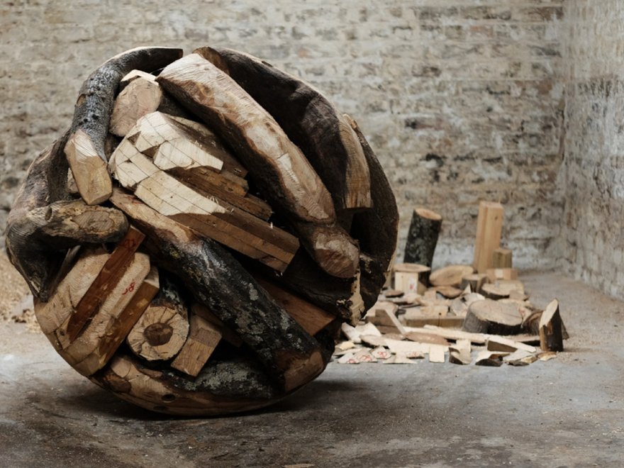 A large, spherical sculpture made of various pieces of cut wood, resembling a giant ball of firewood, sits on a concrete floor in a room with brick walls.