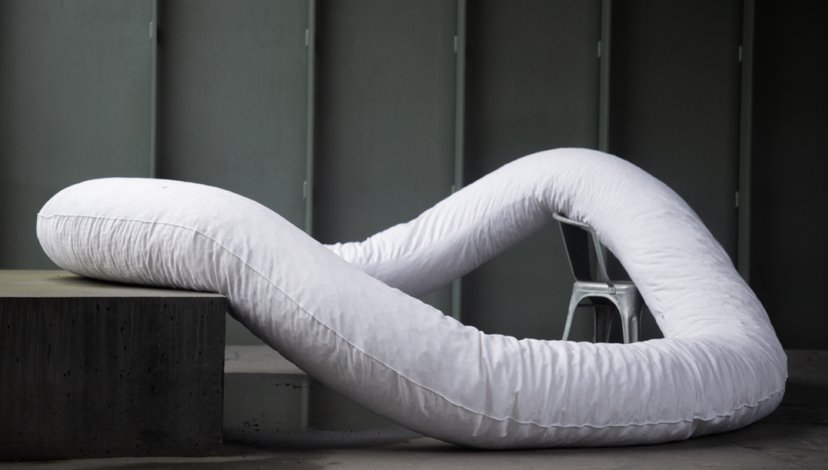 A long, white, flexible body roll cushion or pillow, possibly part of a postural sleep system, shaped into a serpentine loop and resting on a grey block and the floor against a grey wall, with a white chair partially visible in the background.