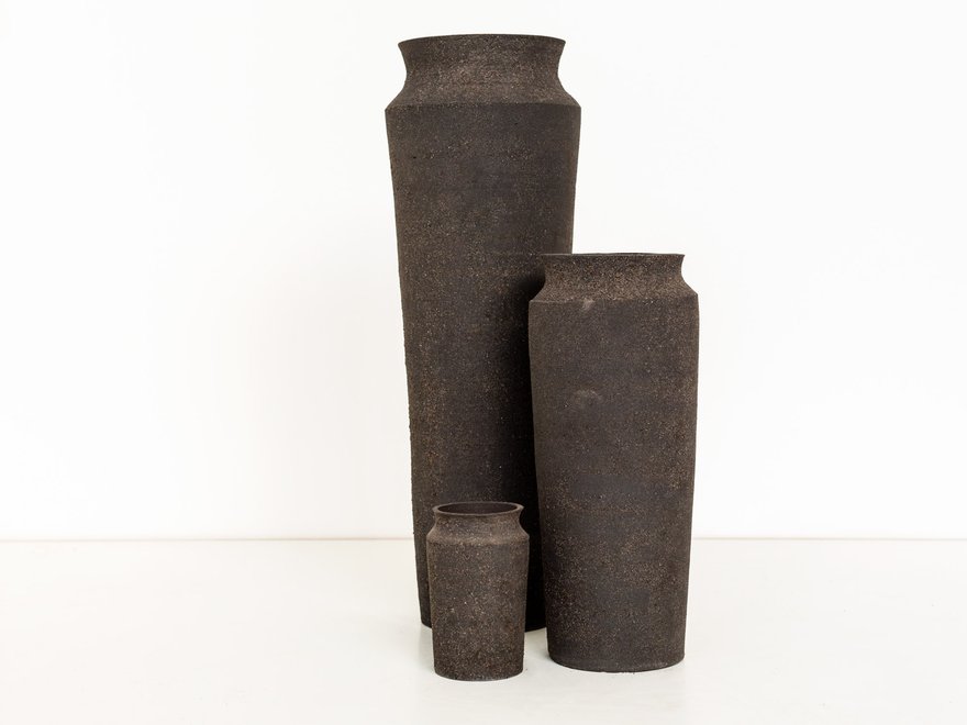 Three dark, textured, vase-like vessels of varying heights stand against a plain white background.