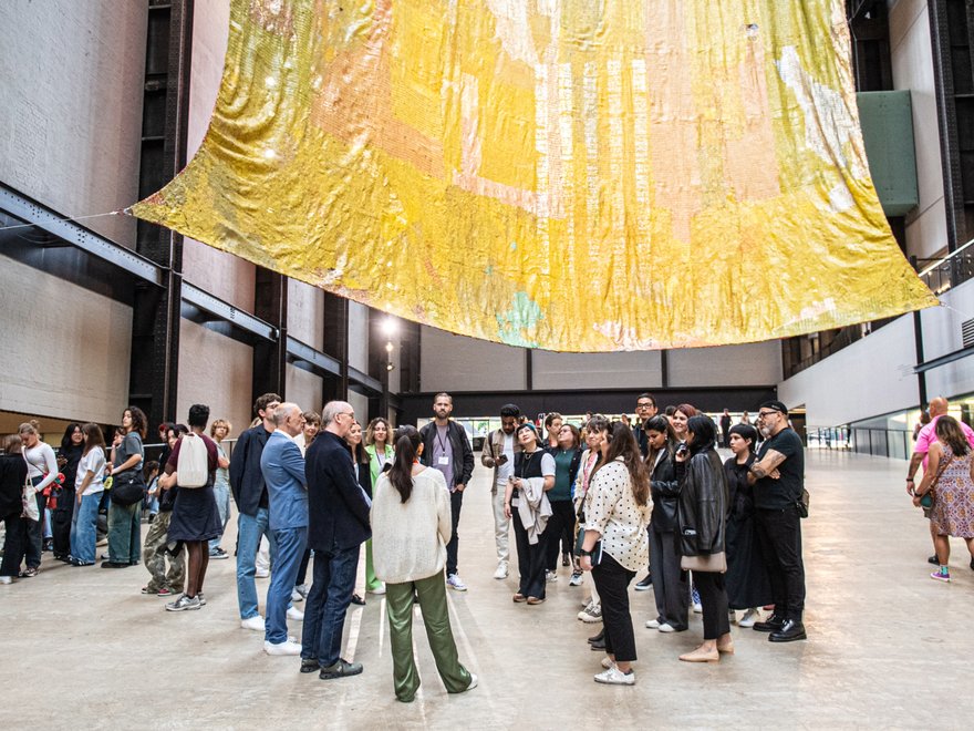 A group of people stand in a large gallery space with a textile hanging above