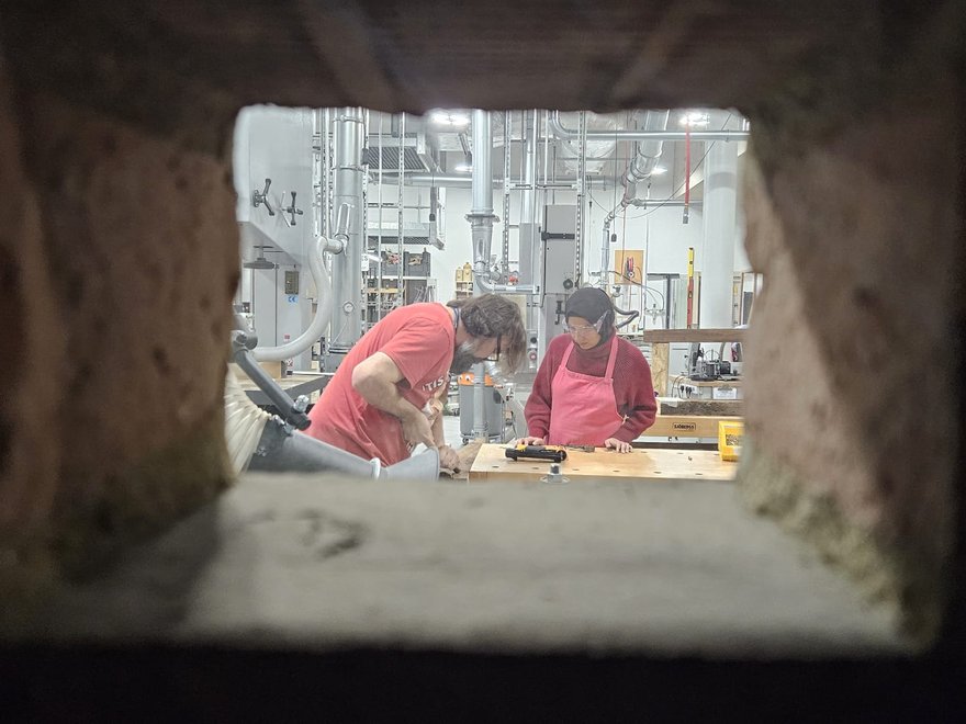 A man and a woman stand at a workbench, photo taken through keyhole