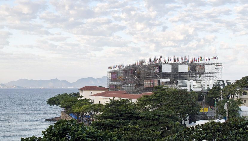 A large, multi-story structure resembling a building or pavilion, surrounded by scaffolding and topped with numerous flags, stands prominently on a coastline next to the ocean.