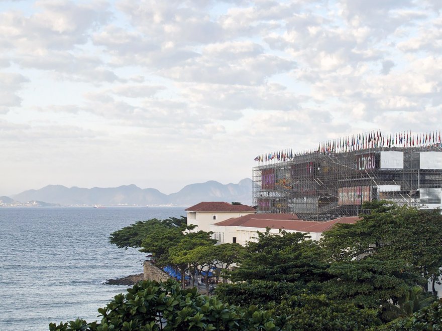 A large, multi-story structure resembling a building or pavilion, surrounded by scaffolding and topped with numerous flags, stands prominently on a coastline next to the ocean.