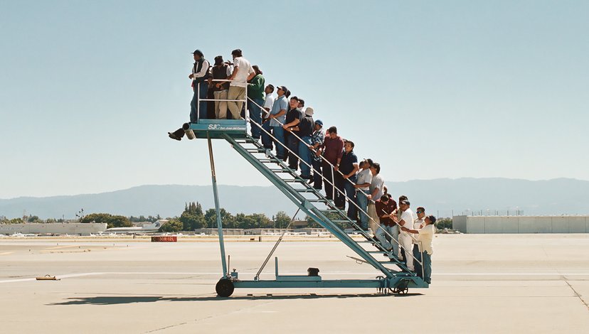 A group of people stands on a mobile airport boarding staircase on an airfield under a clear sky.