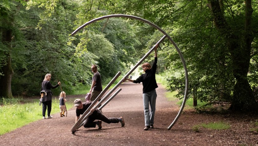 Four individuals are interacting with a large, curved metal sculpture in a park-like setting with trees and a paved path