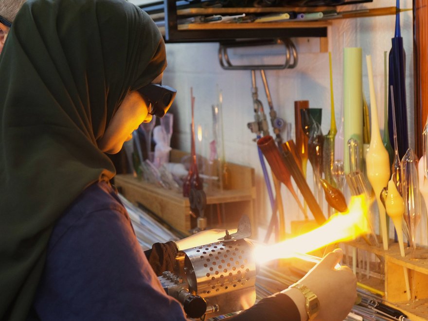 A young girl uses a blow torch to blow coloured glass