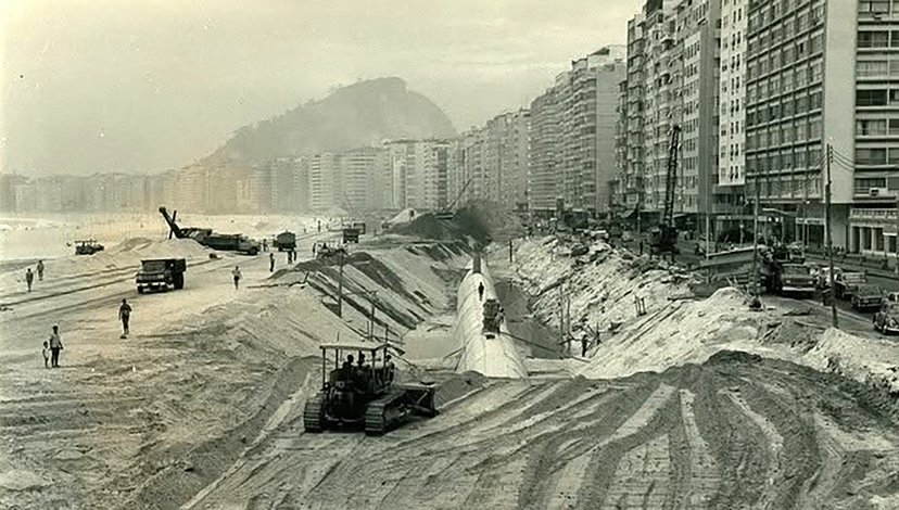 A black and white photo showing construction work on a wide, sandy beach lined with tall buildings.