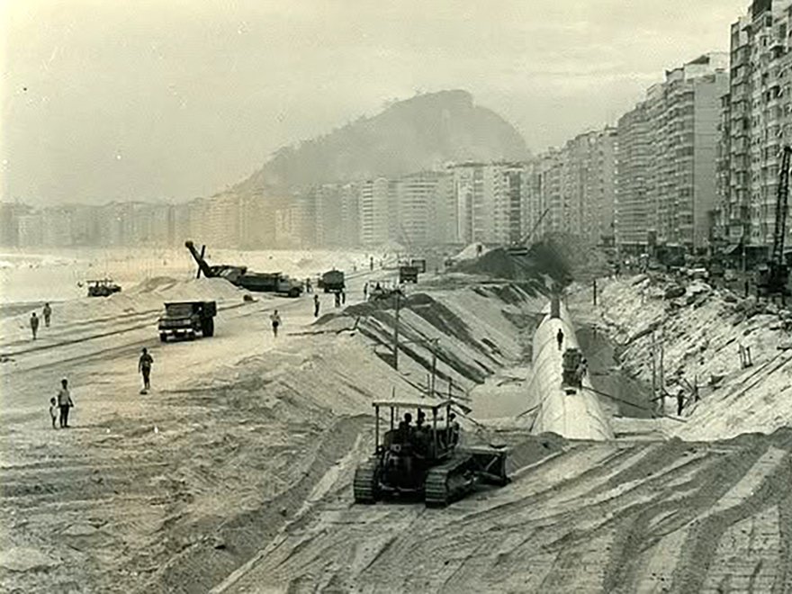 A black and white photo showing construction work on a wide, sandy beach lined with tall buildings.