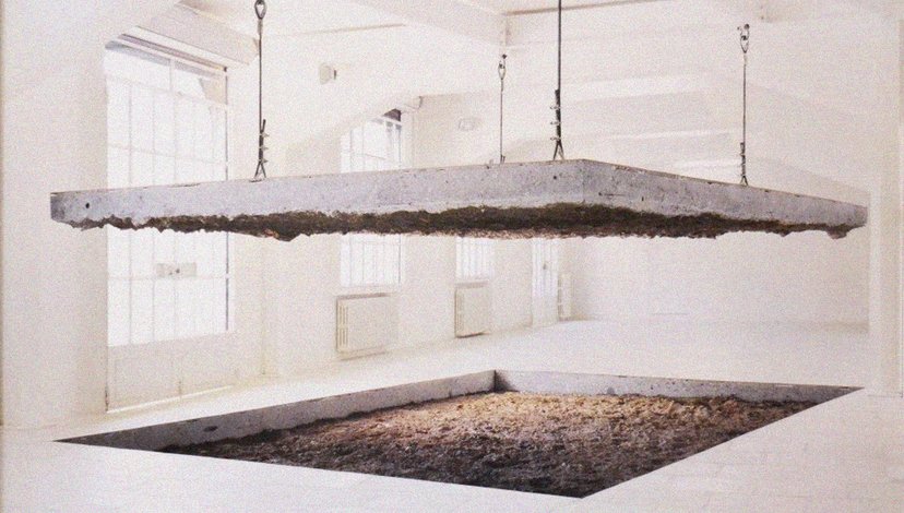 A large, flat concrete slab suspended by chains above a square pit filled with dirt or sand in a white, minimalist gallery space.