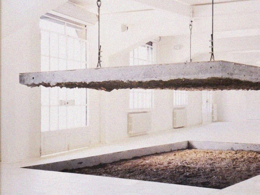 A large, flat concrete slab suspended by chains above a square pit filled with dirt or sand in a white, minimalist gallery space.