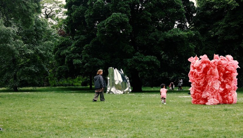 Two children walk across a grassy field in a park, with a large, abstract pink sculpture on the right and a silvery, geometric sculpture in the middle distance. Tall green trees form a backdrop.