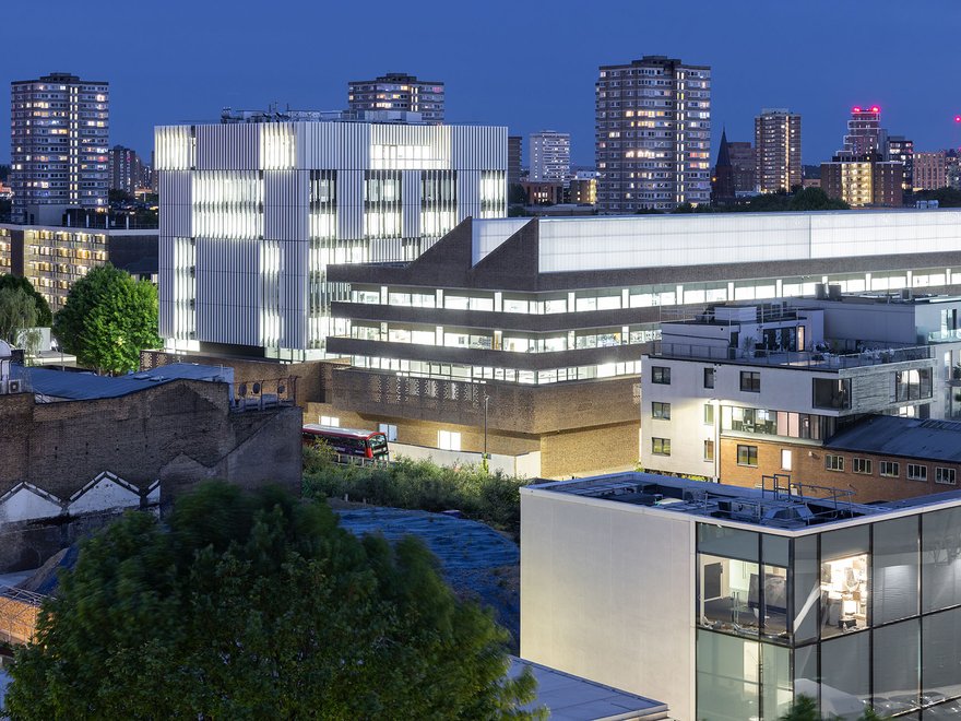 The new RCA Battersea campus at night, lit from within, against an urban London skyline.