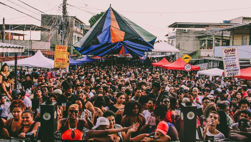 A crowded outdoor street festival or party, likely a 'baile funk' in a favela, with a large, colorful tent in the center and smaller tents on the sides, surrounded by a dense crowd of people.