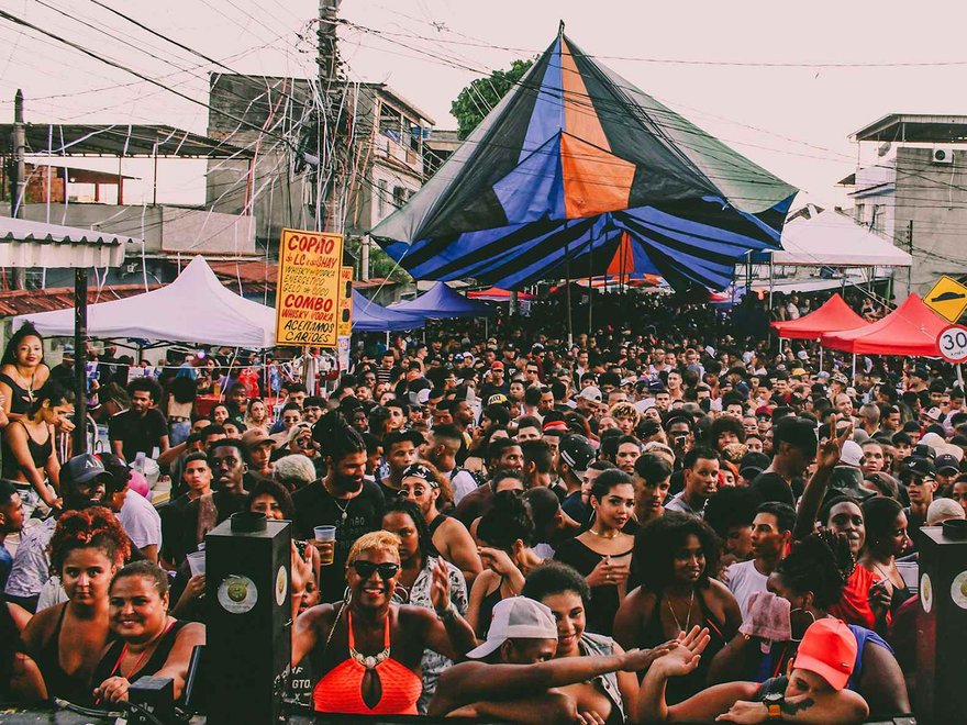A crowded outdoor street festival or party, likely a 'baile funk' in a favela, with a large, colorful tent in the center and smaller tents on the sides, surrounded by a dense crowd of people.