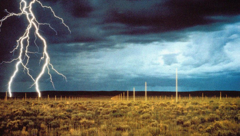 Hundreds of tall, polished stainless steel poles arranged in a grid under a stormy sky with a prominent lightning bolt striking the ground.
