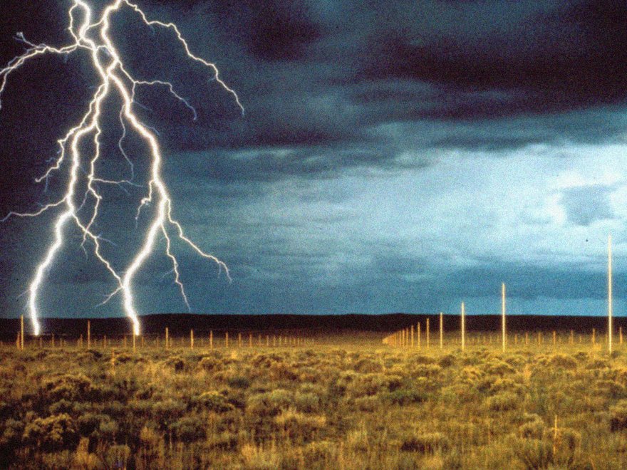 Hundreds of tall, polished stainless steel poles arranged in a grid under a stormy sky with a prominent lightning bolt striking the ground.