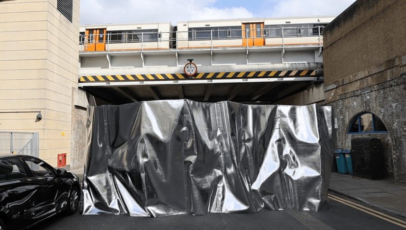 A street view showing a dark car parked on the left, a large silver tarp covering an opening under a railway bridge, and a train visible on the tracks above the bridge.