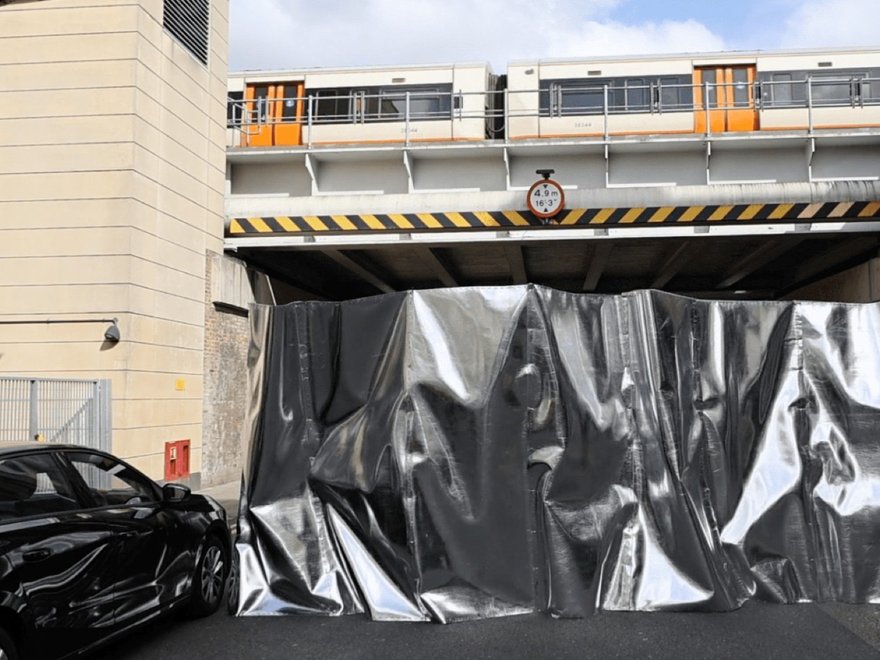 A street view showing a dark car parked on the left, a large silver tarp covering an opening under a railway bridge, and a train visible on the tracks above the bridge.