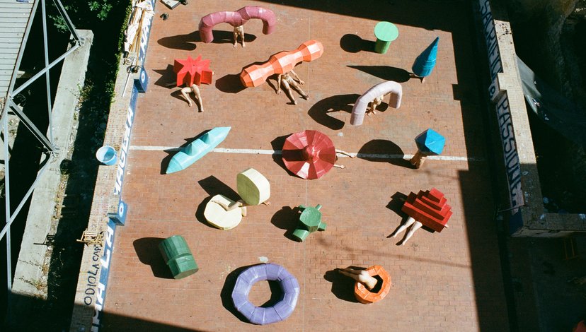 An aerial view of an outdoor installation or exhibition space, possibly a courtyard or paved area, featuring numerous abstract, sculptural objects in various colors and shapes scattered across the ground.
