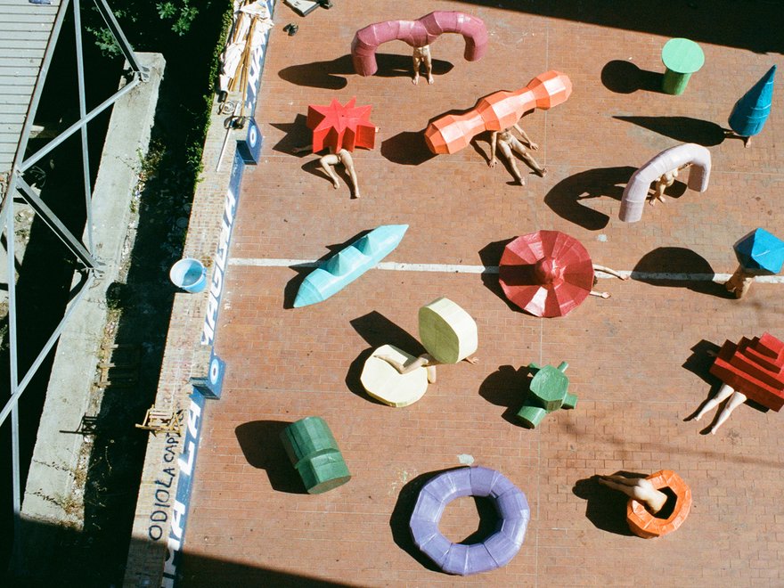 An aerial view of an outdoor installation or exhibition space, possibly a courtyard or paved area, featuring numerous abstract, sculptural objects in various colors and shapes scattered across the ground.