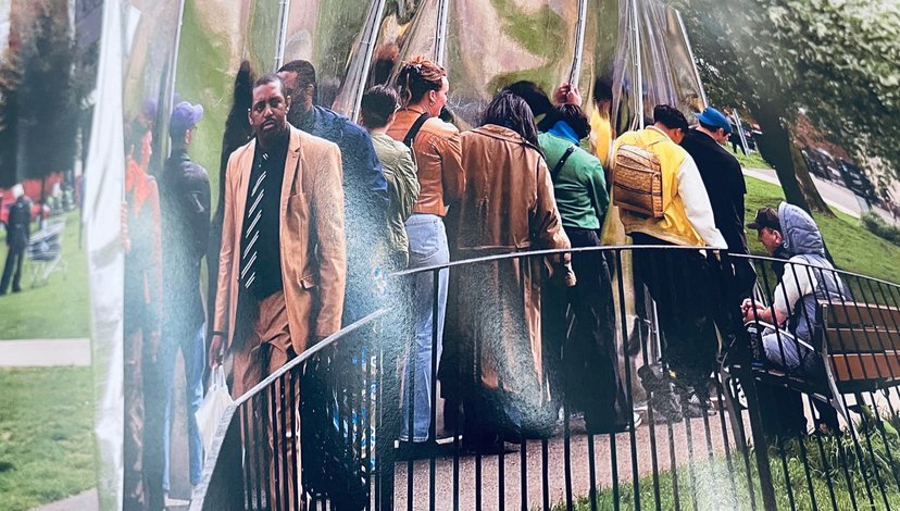 A group of people are gathered on a bridge or walkway, with a man in a tan suit and striped tie prominently in the foreground facing the camera.