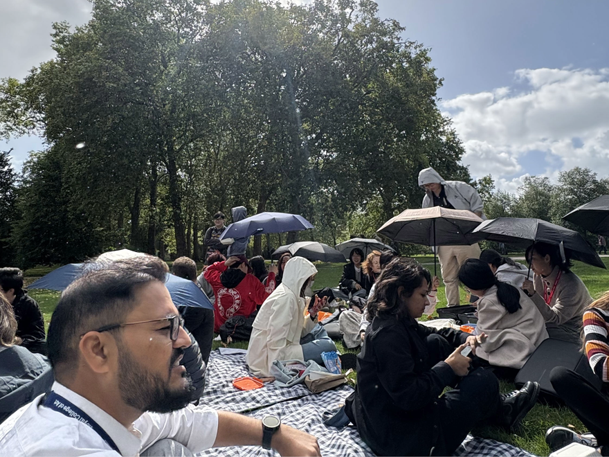 A group of people sit in a sunny park with umbrellas