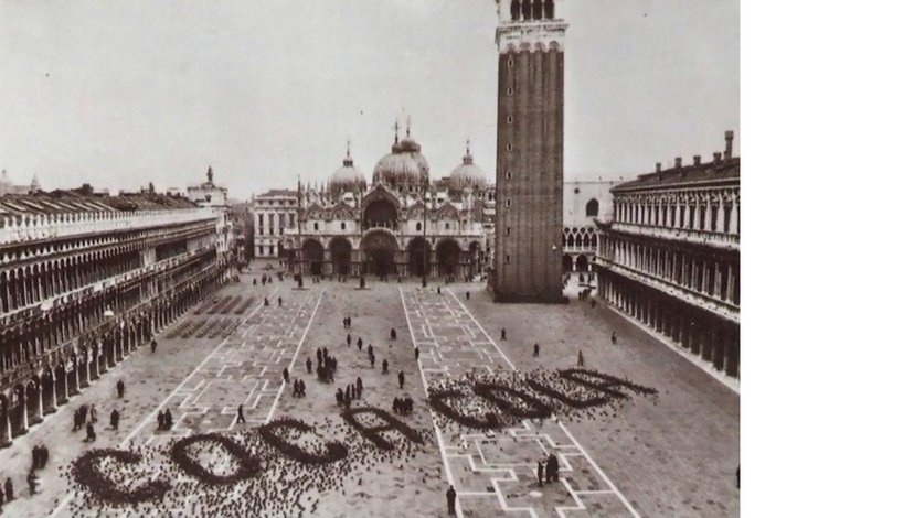 A black and white photograph of St. Mark's Square in Venice, where a large flock of pigeons on the ground forms the word "Coca-Cola" in the foreground.