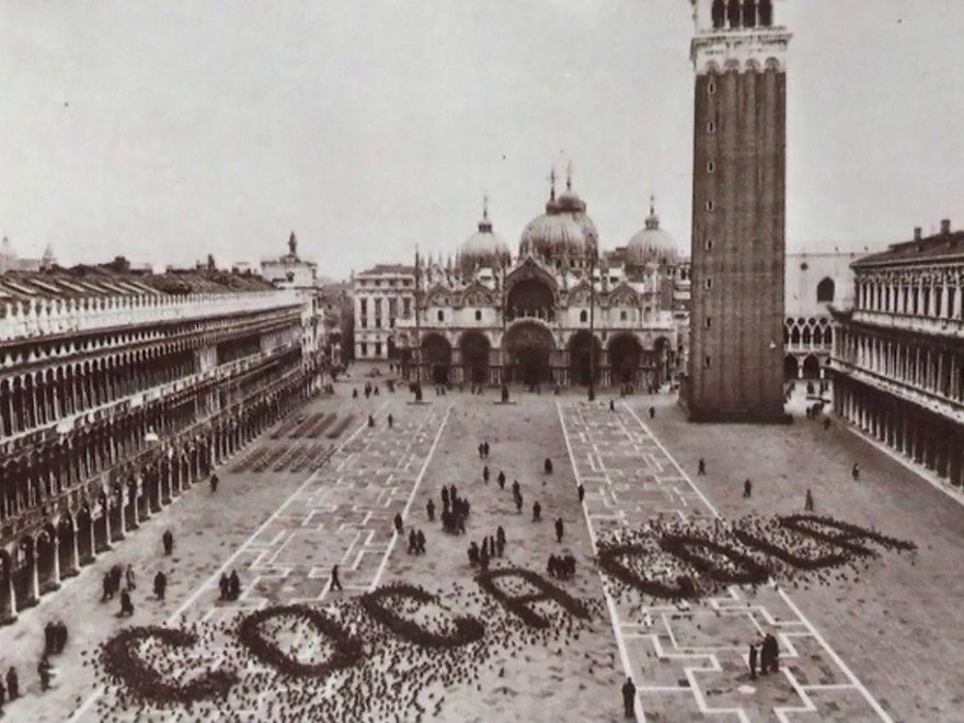 A black and white photograph of St. Mark's Square in Venice, where a large flock of pigeons on the ground forms the word "Coca-Cola" in the foreground.