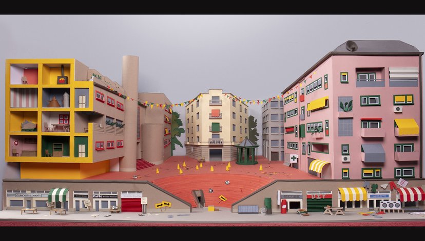 A stylized, colorful diorama of a town square with various buildings, including a large pink apartment building, a yellow multi-story structure with open sections, and a central gazebo in a red-orange square.