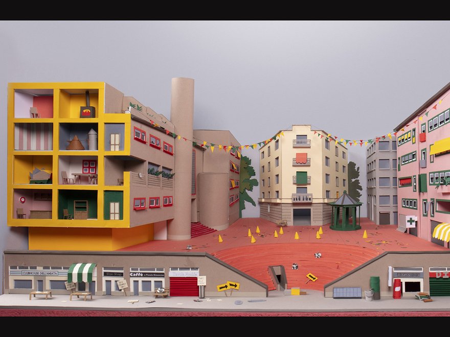 A stylized, colorful diorama of a town square with various buildings, including a large pink apartment building, a yellow multi-story structure with open sections, and a central gazebo in a red-orange square.