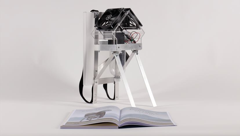 A small, intricate, possibly homemade device resembling a miniature house or structure with exposed wiring and fans sits on a stand, next to an open book on a white surface.
