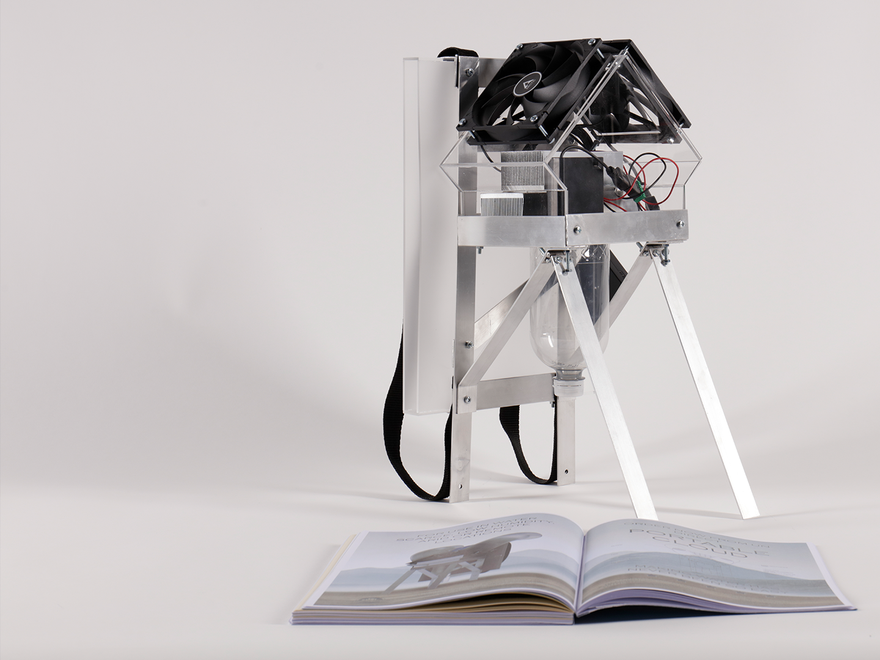 A small, intricate, possibly homemade device resembling a miniature house or structure with exposed wiring and fans sits on a stand, next to an open book on a white surface.