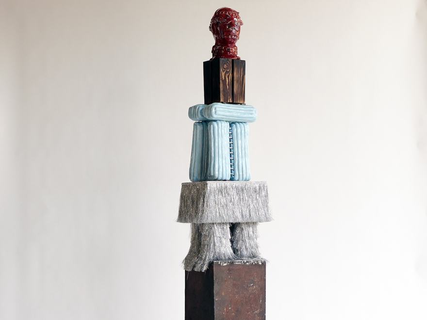 Tall totem sculpture made of stacked textured blocks topped with a red ceramic head.