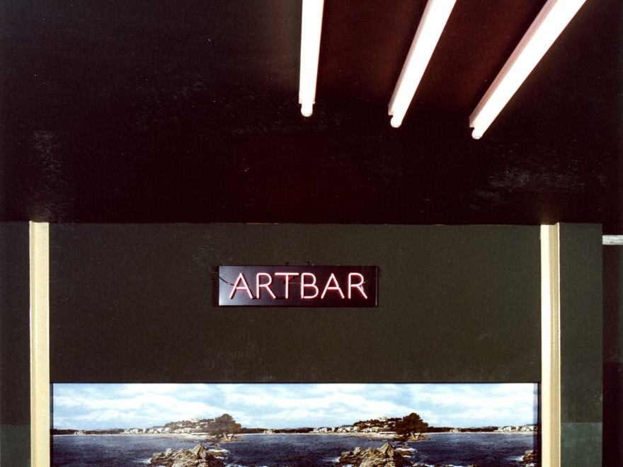 The interior of the ArtBar, a student union bar at the Royal College of Art (RCA) in London. The image features three diagonal pendant light fixtures suspended from a dark ceiling, a neon sign reading "ARTBAR," and a panoramic landscape mural.